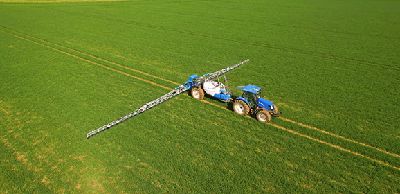 A blue tractor equipped with a long boom sprayer working across a green farmland field, leaving straight crop rows.